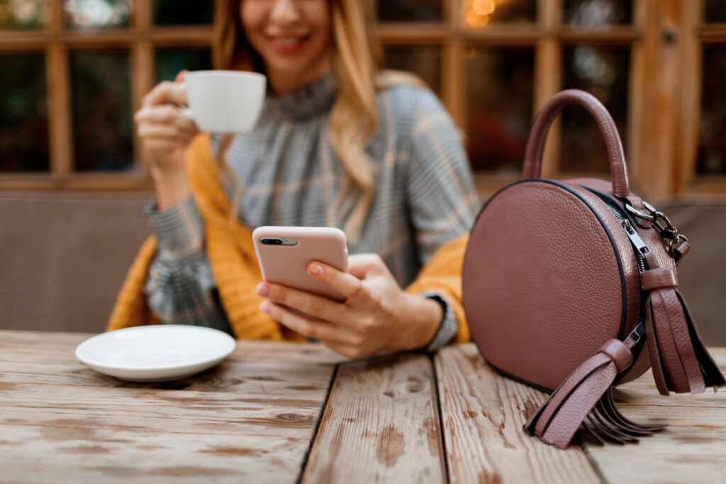 woman using mobile phone texting message drinking coffee stylish bag table wearing grey dress orange plaid enjoying cozy morning cafe